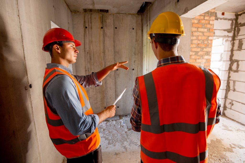 Two civil engineers dressed in orange work vests and helmets walk inside the building under